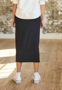 Jupe midi noire en matériau doux et extensible ; présente une coupe droite et des fentes sur les côtés. Associée à des baskets montantes blanches sur un sol en bois.