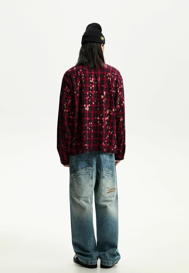 Chemise en flanelle à carreaux rouges et noirs avec des taches de javel, associée à un jean bleu délavé à jambes larges avec un logo brodé "Tiger" sur la poche arrière.