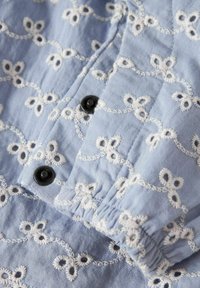 Šviesiai mėlyna medžiaga su baltais siuvinėtais gėlių raštais ir juodais mygtukais; turi raukšlėtą detalę ir tekstūruotą paviršių.