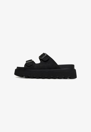 Black suede slide sandals with two adjustable buckles and a thick, textured rubber sole featuring a jagged edge.