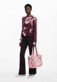 Bolsa de tela rosa con varios bolsillos con cremallera, llevada por una persona que viste un suéter tie-dye marrón y jeans acampanados oscuros. Zapatos blancos.