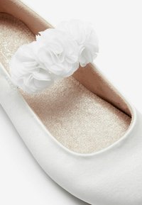 Bailarinas de satén blanco con punta redondeada, con un adorno decorativo de flor blanca y una plantilla brillante color beige.