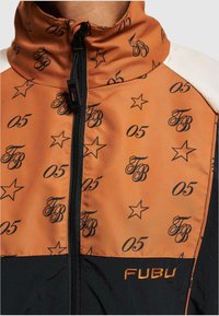 Orange jakke med sort stjerne og skriftmønster, høj krave, lynlås og sort nederste del. Har "FUBU" logo i orange på lommen.