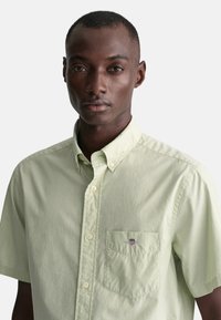 Camisa de botón de manga corta verde claro con un bolsillo en el pecho izquierdo y un logo; hecha de una tela suave y texturizada.