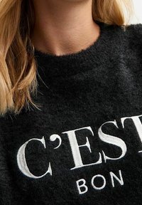 Pull en laine noire duveteux avec un col rond, arborant le texte brodé en blanc "C'EST BON" en évidence sur le devant. Texture douce, sans motifs.