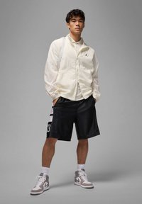 Veste légère blanche avec un logo, superposée sur une chemise blanche. Shorts de sport noirs avec des rayures blanches, associés à des baskets grises et blanches.
