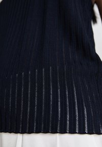 Ναυτικό μπλε πλεκτό τοπ με κάθετες ραβδώσεις και κρόσσια στην άκρη. Το ύφασμα είναι ελαφρύ με υφασμένη εμφάνιση, αναδεικνύοντας το μοτίβο.