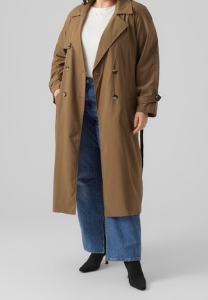 Person, die einen langen braunen zweireihigen Trenchcoat über einem weißen Oberteil, blauen weiter geschnittenen Jeans und schwarzen Absatz-Stiefeletten vor grauem Hintergrund trägt.