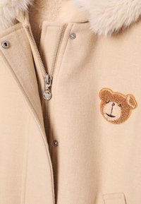 Beżowy płaszcz o miękkiej fakturze, z srebrnym zamkiem błyskawicznym oraz brązową naszywką w kształcie misia na piersi. Kołnierz ozdobiony akcentem z futra.