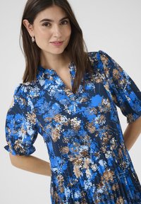 Bloemjurk in marineblauw, met patronen in blauw, bruin en wit. Puffmouwen en een v-hals ontwerp voegen dimensie en textuur toe.