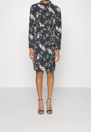 Vrouw draagt een zwarte jurk met lange mouwen met roze en groene bloemen, een ceintuur in de taille en zwarte enkellaarsjes met hoge hakken.