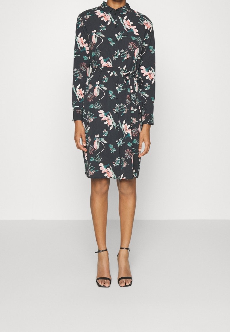 Femme portant une robe noire à manches longues avec des fleurs roses et vertes, taille ceinturée, et des sandales noires à talons hauts avec bride à la cheville.