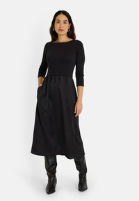Vestido negro con un cuerpo ajustado, mangas de tres cuartos y una falda acampanada fluida con bolsillos. Combinado con botas negras hasta la rodilla.