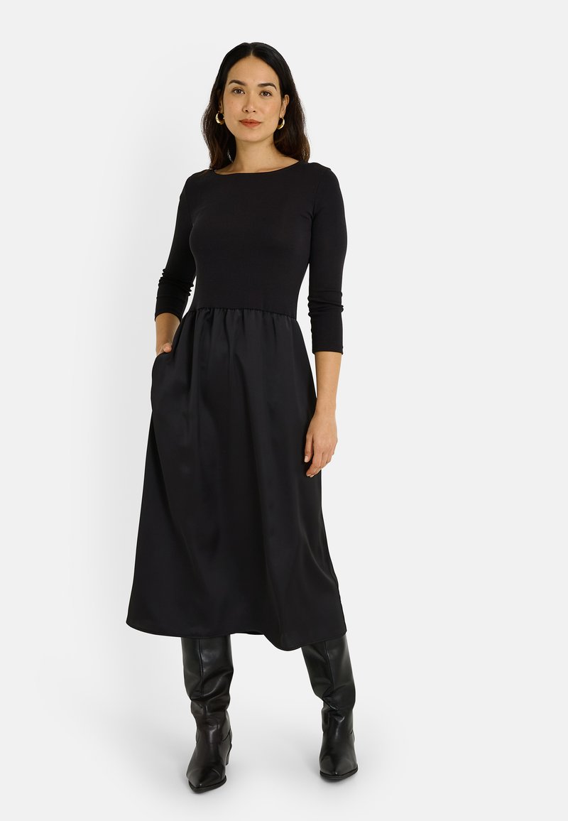 Vestido negro con un cuerpo ajustado, mangas de tres cuartos y una falda acampanada fluida con bolsillos. Combinado con botas negras hasta la rodilla.