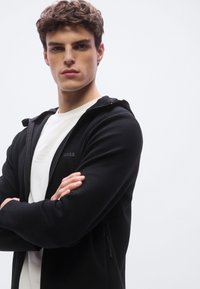 Sudadera negra con cremallera presenta un diseño elegante con un bolsillo delantero, combinada con una camiseta blanca de cuello redondo que muestra un logo de la marca sutil.