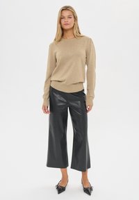 Bež pleteni pulover s dugim rukavima i rebrastim rubom, uparen s sjajnim crnim lažnonatručenim hlačama širokog kroja i crnim cipelama s oštrim vrhom.