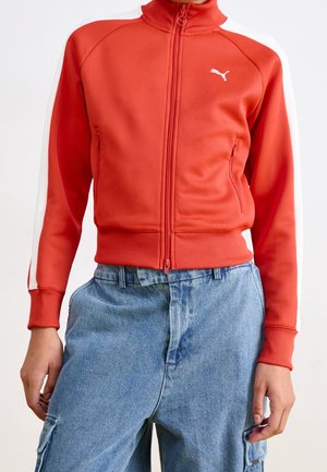 Person trägt eine rote Puma-Jacke mit Reißverschluss, weißen Streifen an den Ärmeln und blaue, hoch taillierte Cargo-Jeans.