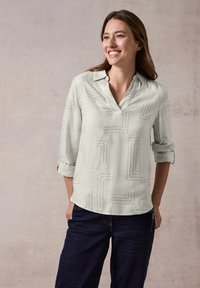 Blusa grigia chiara con scollo a V e maniche arrotolate, caratterizzata da linee geometriche nere. Indossata con jeans scuri, su uno sfondo neutro.
