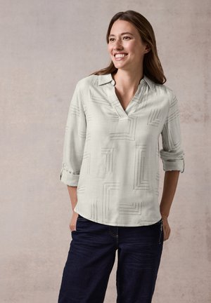 Lichtgrijze blouse met een V-hals en opgerolde mouwen, met zwarte geometrische lijnen. Gepaard met donkere jeans, tegen een neutrale achtergrond.