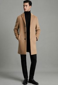 Palton din lână de cămilă cu reveruri ondulate, trei butoane negre și buzunare laterale, purtat cu un pulover negru cu guler și pantaloni negri drepti.