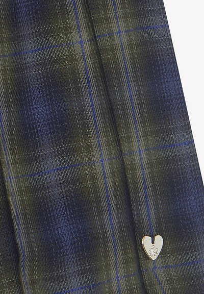 Gros plan sur un tissu à carreaux bleu et vert avec un petit charm en forme de cœur en argent accroché près du coin inférieur droit.