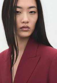Vínový blejzr s ostrými klopami, hladkou tkaninou a na míru šitým střihem. Dlouhé rovné černé vlasy částečně zakrývají tvář modelky.