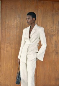 Blazer blanc texturé et pantalon assorti, avec des manches longues et une taille ceinturée. Portant un sac noir. Fond en bois.