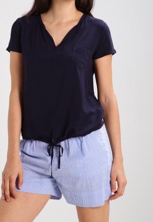 Vrouw draagt een marineblauw T-shirt met korte mouwen en V-hals met borstzak, gecombineerd met lichtblauwe en wit gestreepte shorts met trekkoord.