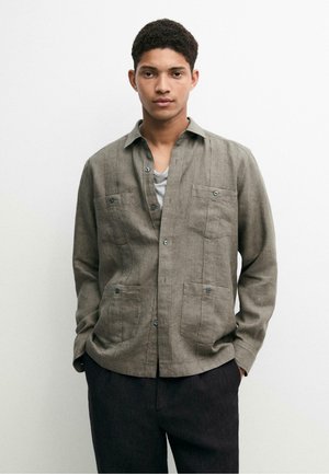 Jeune homme portant une chemise olive ouverte sur un t-shirt gris, les mains dans les poches d'un pantalon noir, debout devant un fond blanc.