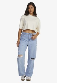 Cropped cream t-shirt with embossed logo, paired with light blue distressed jeans featuring large ripped accents and a relaxed fit.