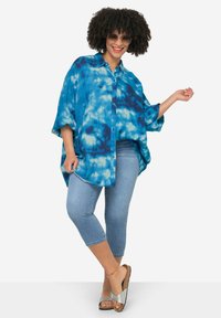 Maglietta blu con stampa tie-dye, dal fit oversize, con chiusura a bottoni e maniche larghe, abbinata a jeans capri blu chiaro e sandali argentati.