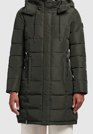 Femme portant un manteau matelassé vert foncé mi-long avec capuche et poches zippées, debout les mains détendues le long du corps.