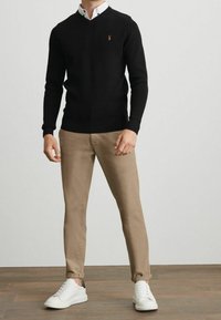 Pull noir à col en V avec texture côtelée et logo orange, associé à un chino beige ajusté et des baskets blanches avec un accent noir.