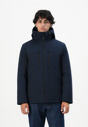 Young man wearing a navy blue hooded jacket zipped up, standing against a plain white background.