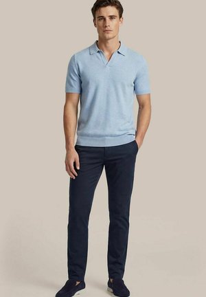 Man staat met één hand in de zak, gekleed in een lichtblauw poloshirt met korte mouwen en donkerblauwe broek met donkere schoenen, tegen een beige achtergrond.