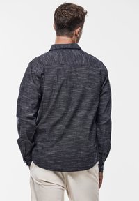 Tmavá košile s dlouhými rukávy a vzorem, která má texturovanou látku a zakřivený lem. Zadní pohled ukazuje standardní design límce.