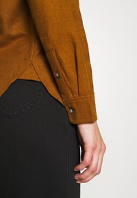 Gros plan sur la main d’une personne posée à son côté, portant une chemise marron à manches longues et un pantalon noir avec des coutures visibles sur la poche arrière.