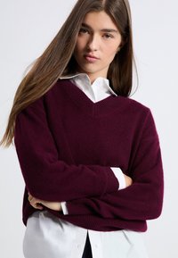 Bordowy, oversize sweter z dekoltem w kształcie litery V, uszyty z miękkiego materiału, w połączeniu z białą koszulą z kołnierzykiem. Sweter ma ściągacze przy mankietach i u dołu.