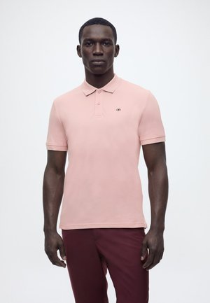 Mężczyzna ubrany w jasnoróżową koszulkę polo z krótkim rękawem i ciemnoczerwone spodnie, stojący na jednolitym jasnym tle.