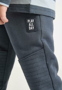 Primo piano dei pantaloni scuri grigi di un bambino con toppe cucite sulle ginocchia e un'etichetta "GIOCA TUTTO IL GIORNO" vicino all'anca, con una mano parzialmente visibile.