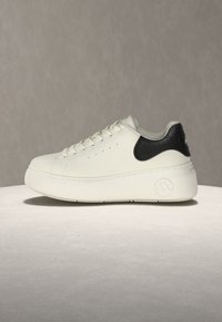 Tênis branco de couro com calcanhar preto texturizado. Apresenta uma sola plataforma grossa e biqueira arredondada, com perfurações visíveis na lateral.