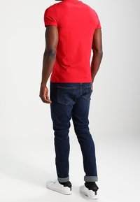 Rotes Kurzarmhemd kombiniert mit dunkelblauen Jeans. Die Jeans haben eine leicht taillierte Passform und gerollte Säume. Weiße Sneakers runden das Outfit ab.