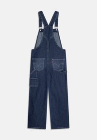 Džínové lacláče v tmavě modré barvě s nastavitelnými popruhy, širokými nohavicemi, dvěma předními kapsami a boční kapsou na nářadí. Detaily kontrastního šití.