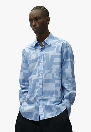 Uomo che indossa una camicia azzurra chiara con grandi motivi testuali chiari, in piedi con le mani nelle tasche su sfondo bianco.