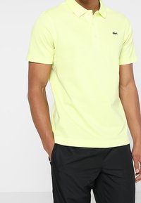 Limetkově zelené polo tričko s zapínacím límečkem a krátkými rukávy, na levé straně hrudi má malé černé logo. Pod ním jsou černé kalhoty.