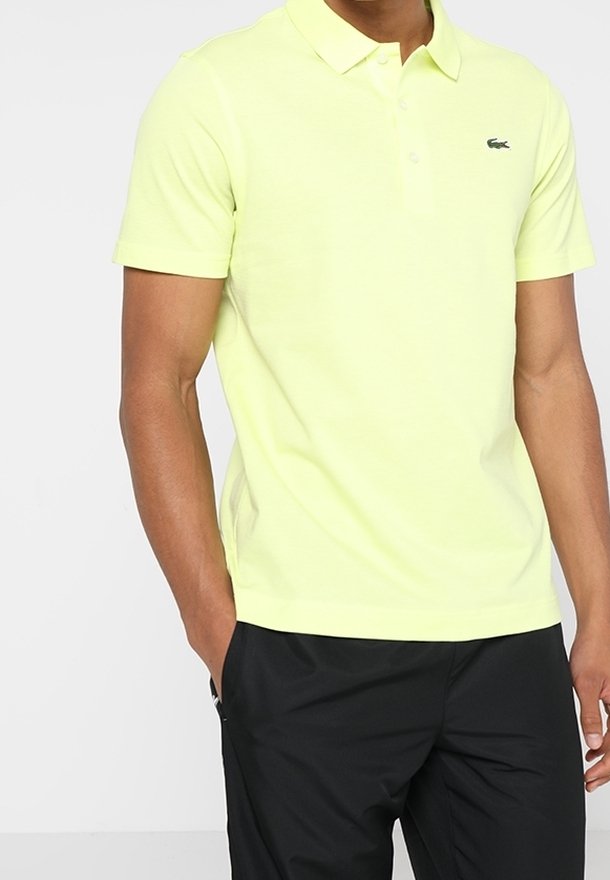 Limetkově zelené polo tričko s zapínacím límečkem a krátkými rukávy, na levé straně hrudi má malé černé logo. Pod ním jsou černé kalhoty.