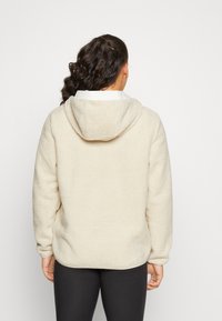 Sudadera de felpa beige con una superficie texturizada, capucha ajustable y mangas largas. El dobladillo es ligeramente curvado, proporcionando un ajuste relajado.