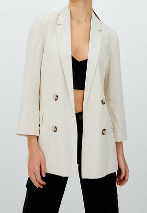 Frau trägt einen beigen zweireihigen Blazer über einem schwarzen Crop-Top und schwarzen Hosen und steht vor einem einfarbigen Hintergrund.