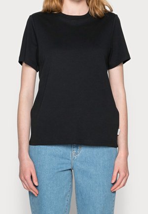 Woman wearing a plain black short-sleeve t-shirt and light blue jeans, standing against a plain light background.