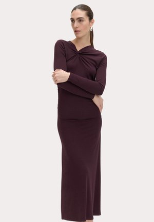 Frau mit langen dunklen Haaren, die ein tailliertes, langärmeliges, burgunderfarbenes Kleid mit verdrehtem Detail am Ausschnitt trägt, steht mit verschränkten Armen.